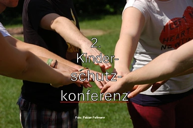 Mehrere Personen stehen auf einer Wiese im Kreis und strecken ihre Arme zur Mitte, wo ihre Hände übereinanderliegen. Über dem Bild steht der Text „2. Kinderschutzkonferenz“, unten ist der Fotohinweis „Foto: Fabian Ellmann“ eingeblendet.